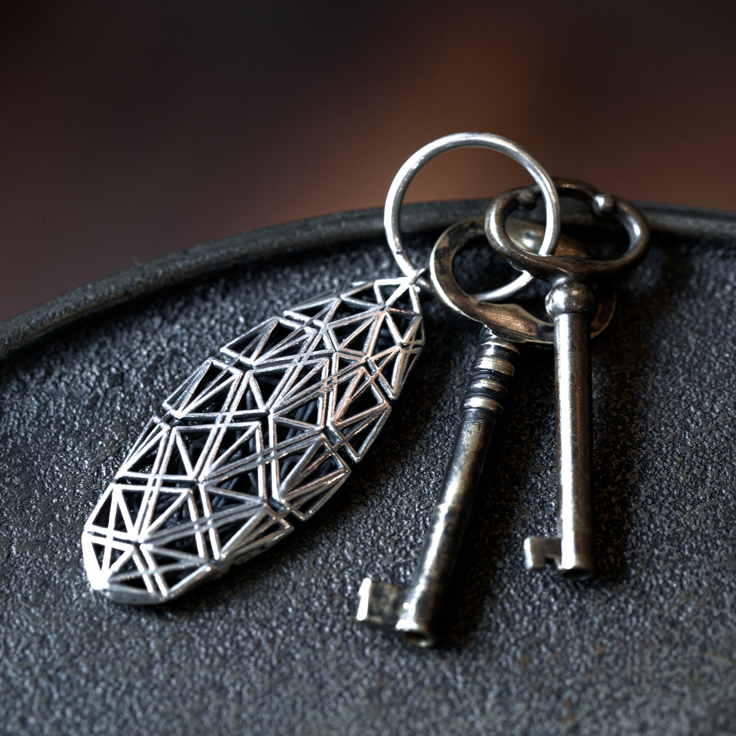 A sterling silver angular lattice designer keychain with mirror-polished surfaces, offset by contrast-oxidized matte black detailing, stands out against a dark tabletop.