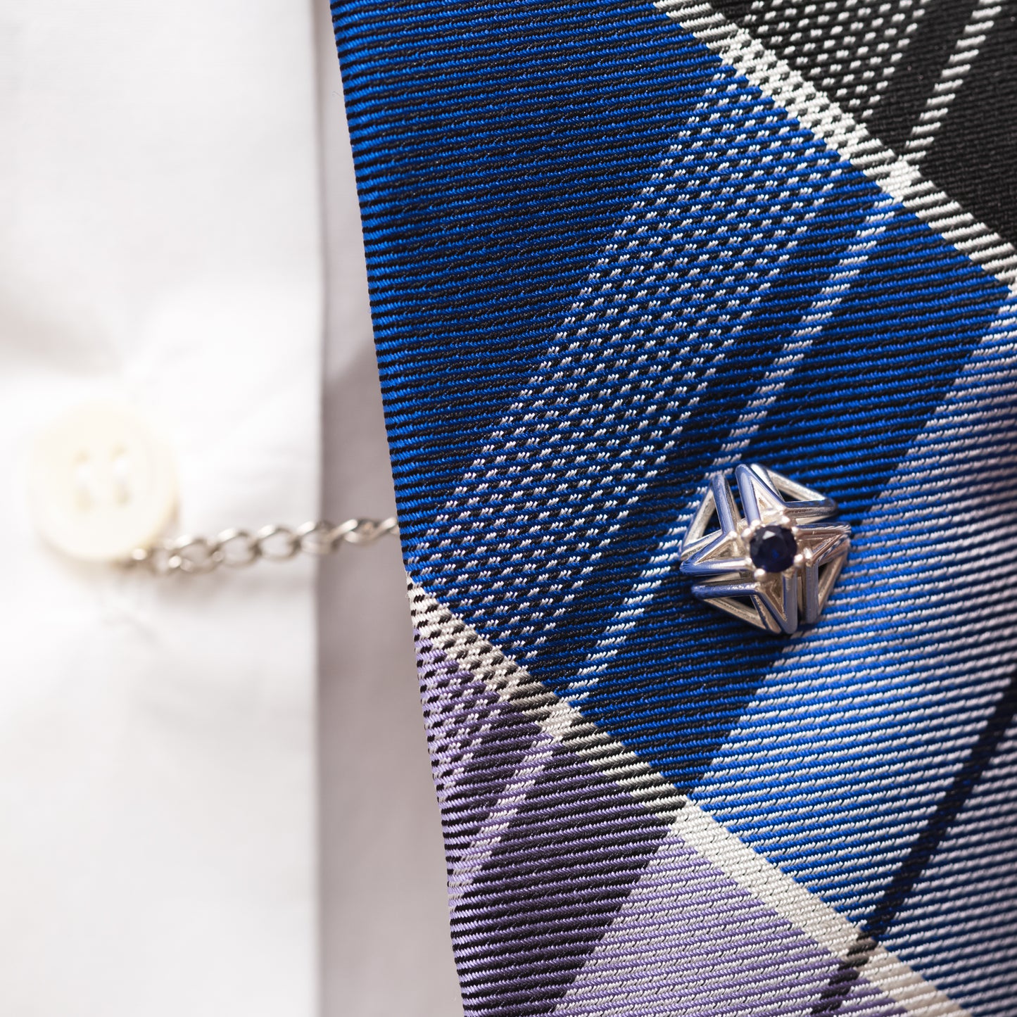 Blue sapphire and solid sterling silver tie tack worn with a blue plaid tie and white button down shirt. The tie tack’s security chain keeps the tie centered over the button placket, a detail that helps make this unisex accessory fit for the modern dandy.