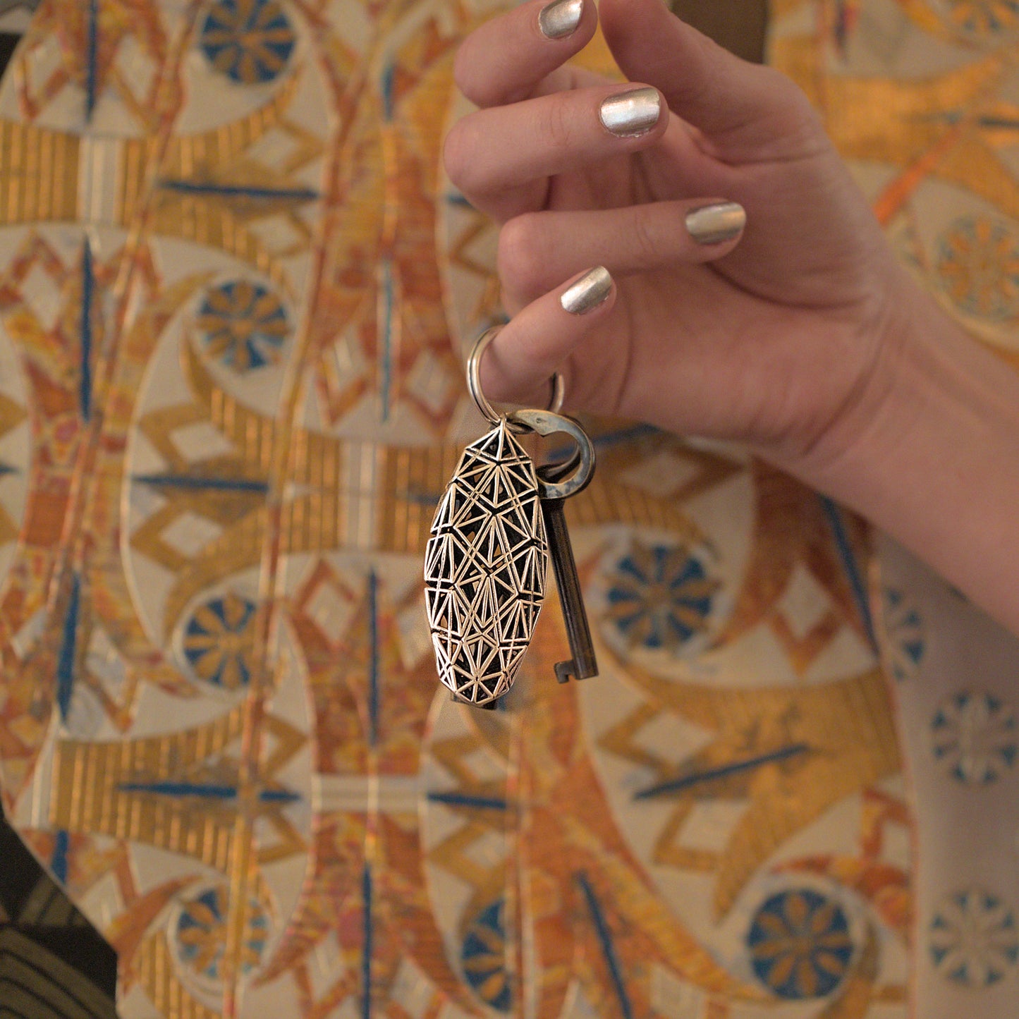 A sterling silver keychain defined by precise architectural geometry hangs from a woman's pinky finger. The key ring is large enough to accommodate multiple keys. The bold design stands out against any backdrop. 