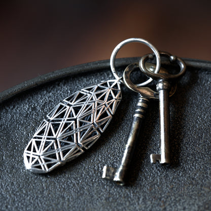 A sterling silver angular lattice designer keychain with mirror-polished surfaces, offset by contrast-oxidized matte black detailing, stands out against a dark tabletop.